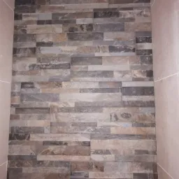 Ściana wyłożona podłużnymi płytkami imitującymi kamień w odcieniach szarości i beżu, widok z bliska na fragment ściany z widocznym fragmentem sufitu i ścian bocznych.