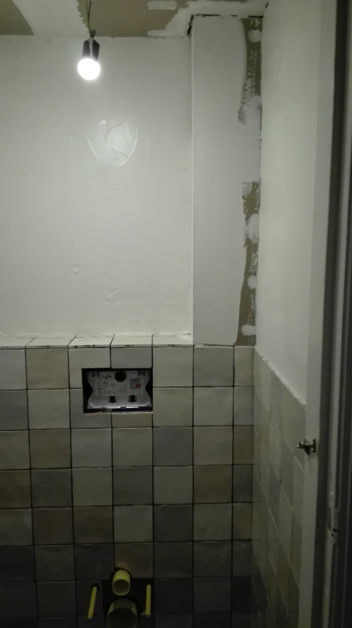 Łazienka w trakcie remontu: częściowo położone szare płytki, odkryta instalacja podtynkowa WC, ściany z śladami szpachlowania i świeżo pomalowane fragmenty.