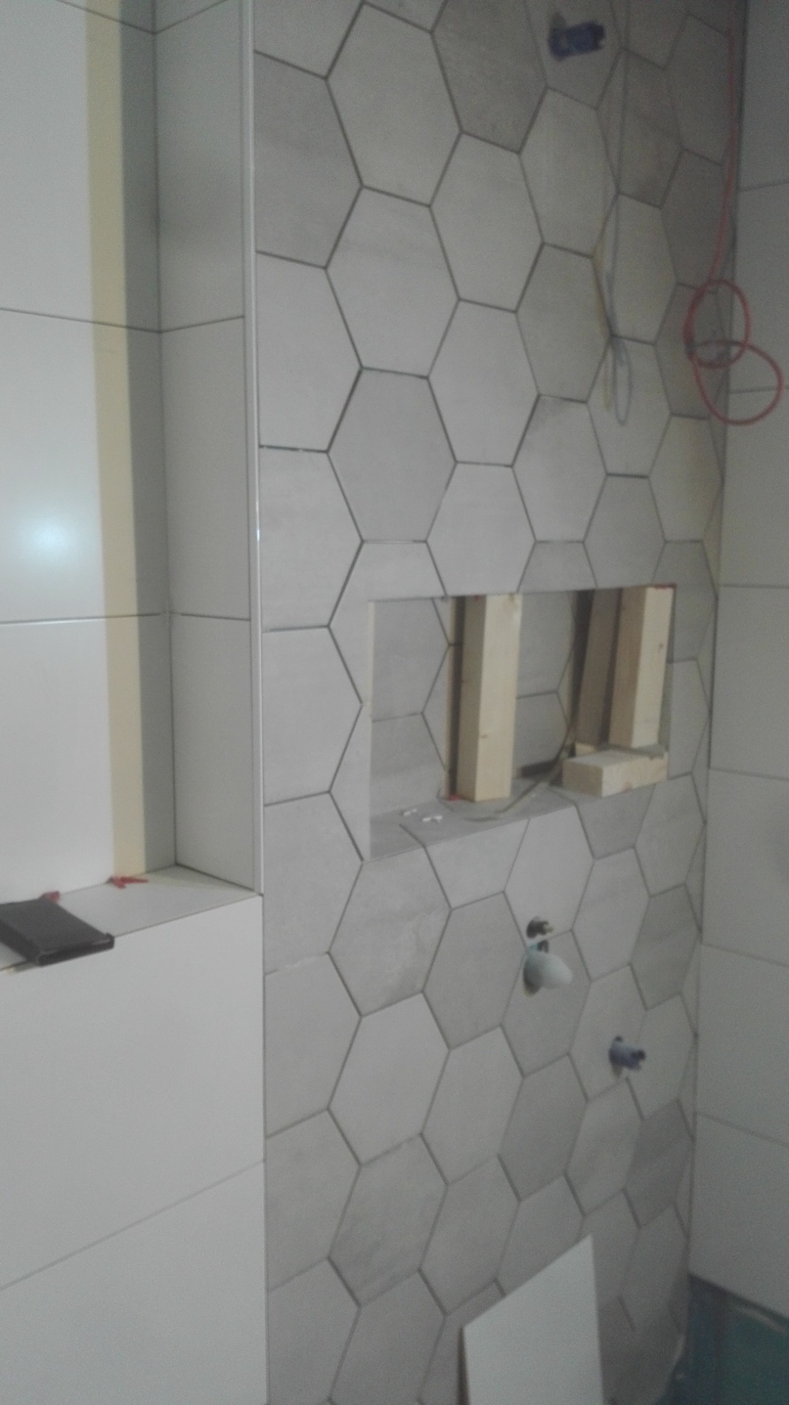 Ściana łazienki wyłożona szarymi, heksagonalnymi płytkami ceramicznymi. Widoczna wnęka w ścianie z drewnianą konstrukcją wewnątrz oraz wystające rury instalacji wodnej. Na półce leży czarny przedmiot.