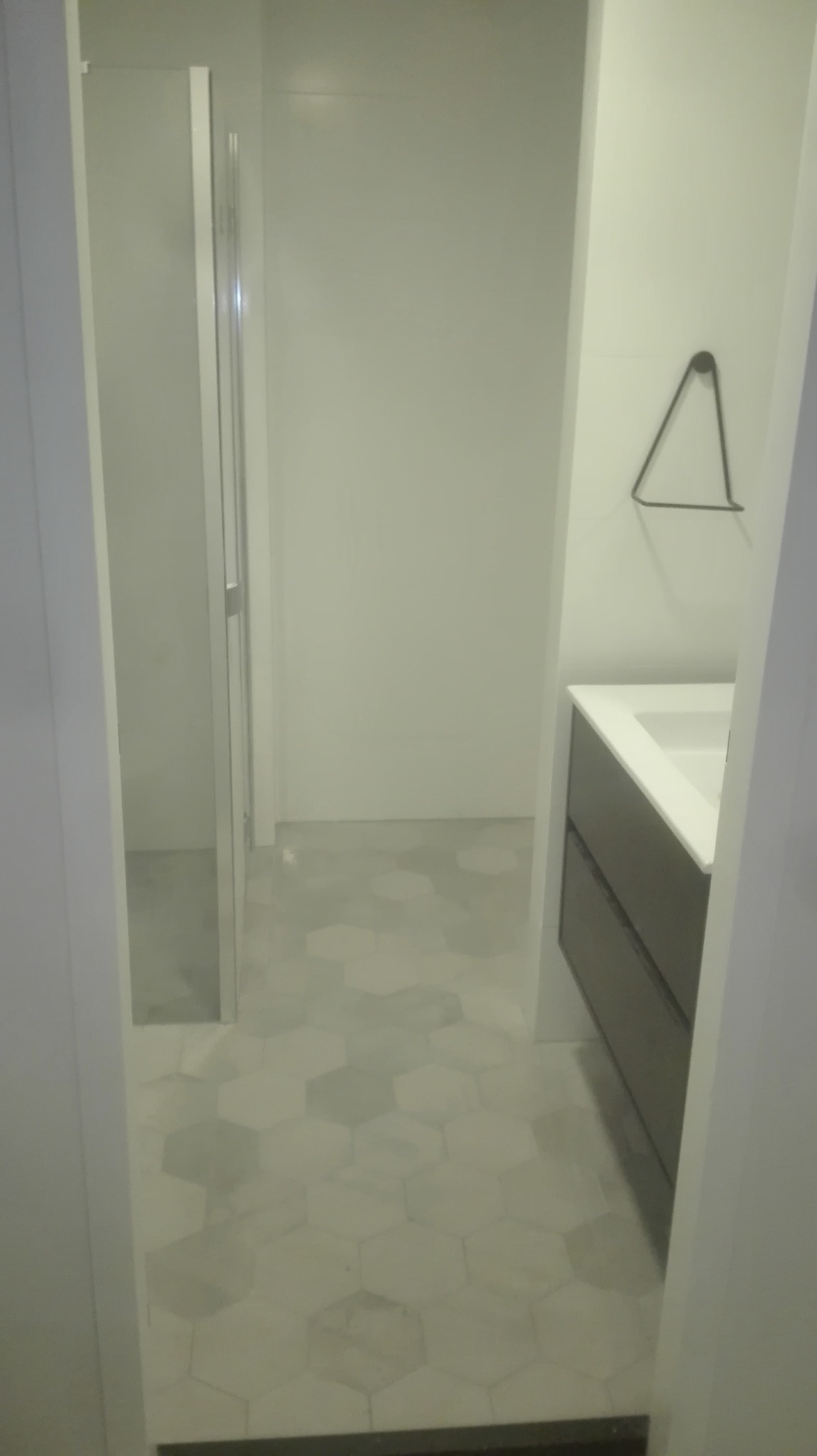 Wykończona łazienka z szarą szafką pod umywalką, kabiną prysznicową i heksagonalną szarą podłogą, widok zza framugi drzwi.