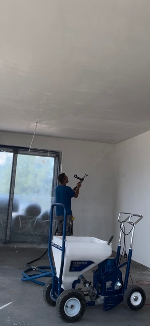 Pracownik rozprowadza masę szpachlową za pomocą agregatu natryskowego Texspray T-Max 506, widoczne surowe ściany i okno.