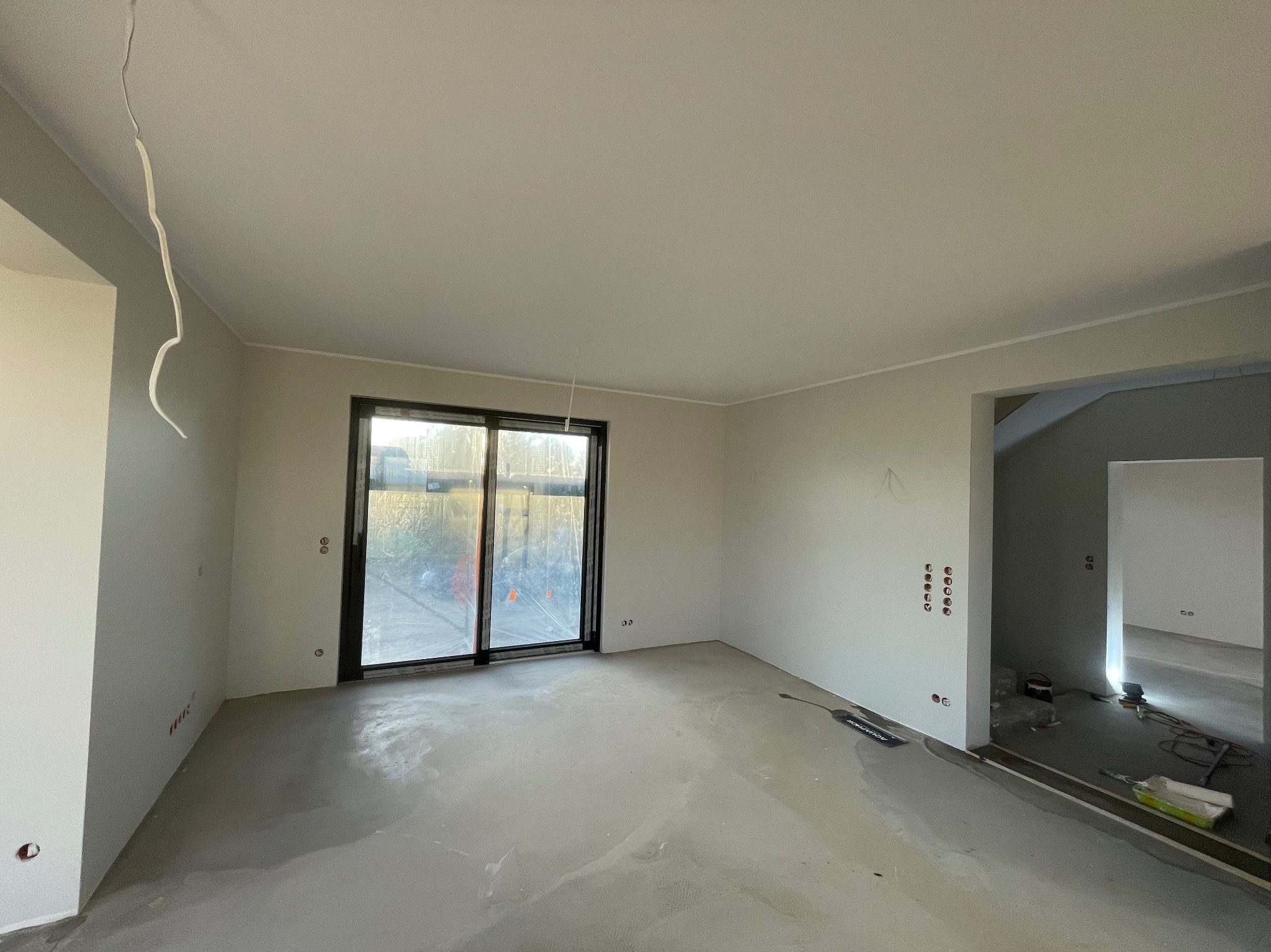 Pokój w stanie deweloperskim z gładzią na ścianach, widoczne gniazdka elektryczne, okno przesuwne z czarnymi ramami, w tle widoczne inne budynki.