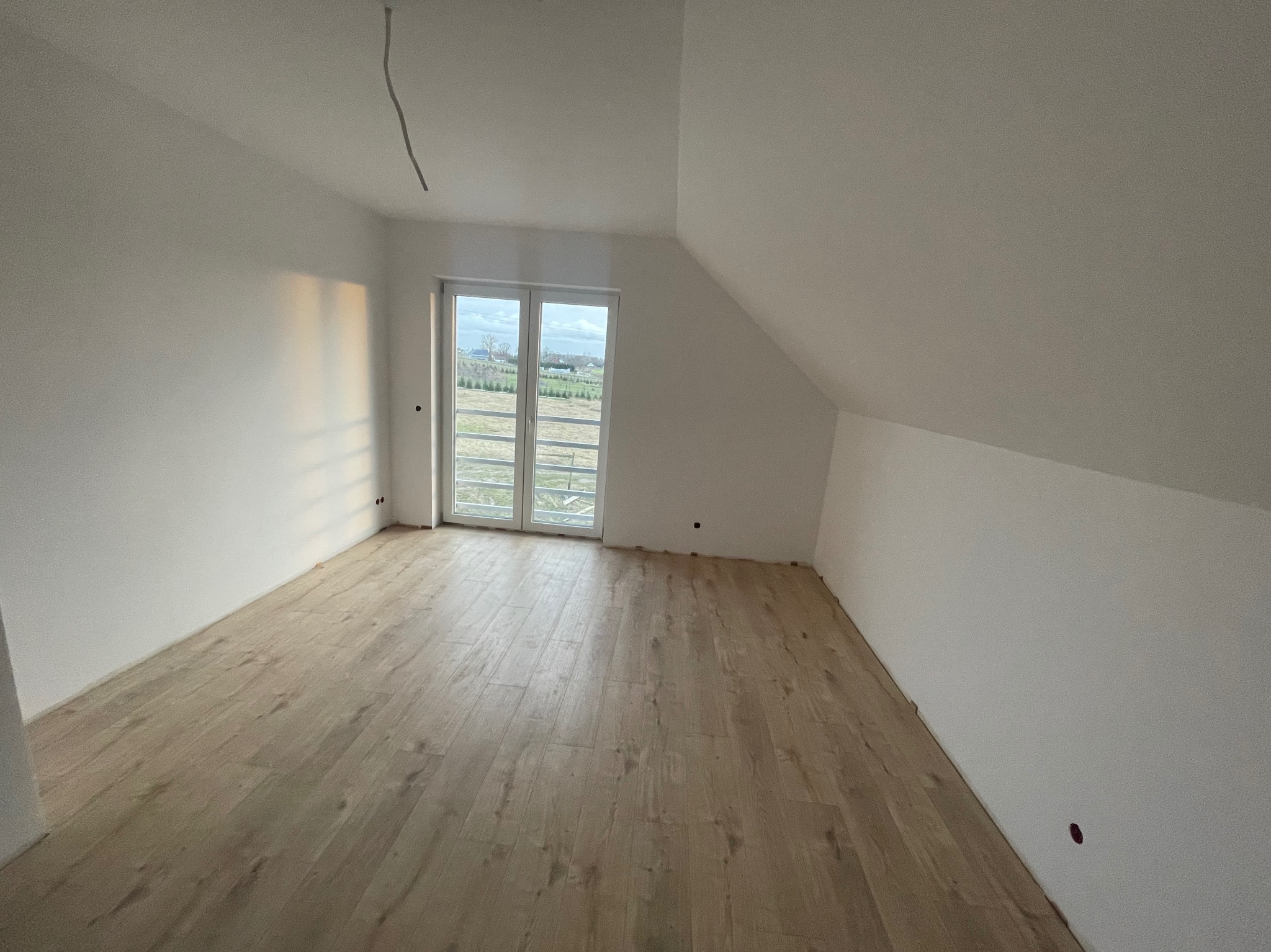 Poddasze z oknem i drewnopodobną podłogą, ściany przygotowane pod malowanie, widoczne niedoskonałości w postaci wystającego kabla i otworu w ścianie.