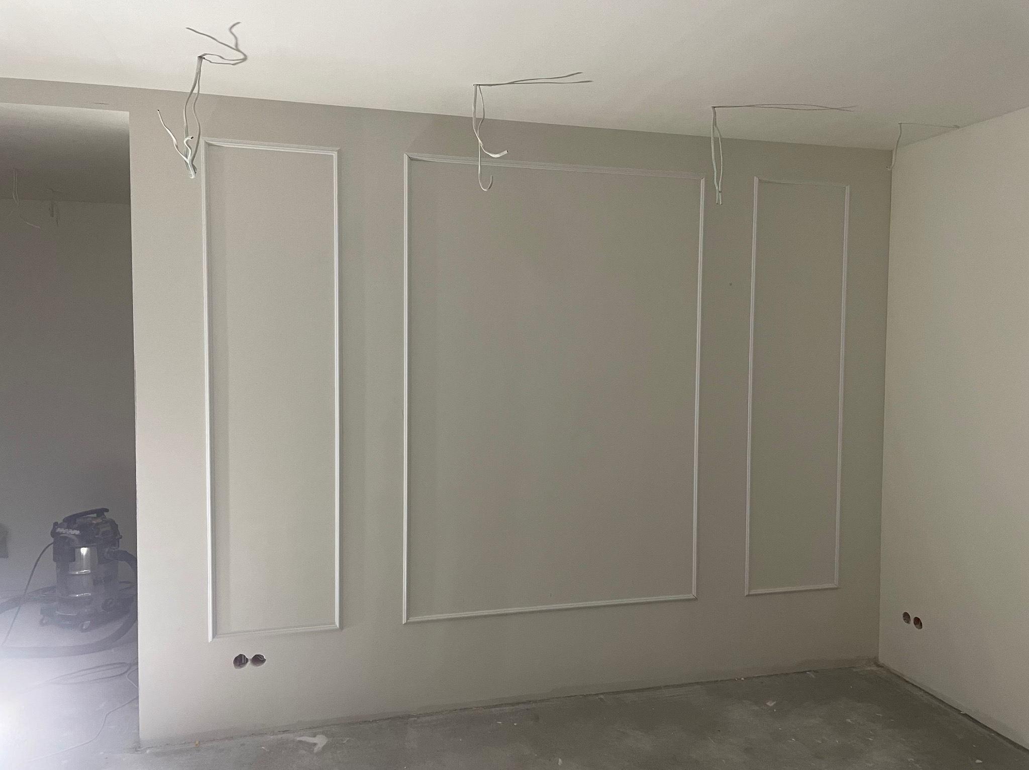 Pokój w trakcie wykańczania, ściany z dekoracyjnymi listwami i wystającymi przewodami elektrycznymi, w tle widoczny odkurzacz przemysłowy.