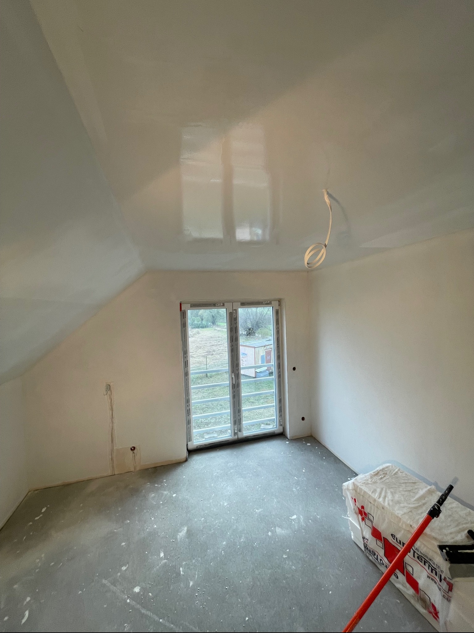 Pokój na poddaszu po nałożeniu gładzi, z widocznymi śladami szpachlowania na podłodze i pakietem materiałów budowlanych w rogu pomieszczenia.