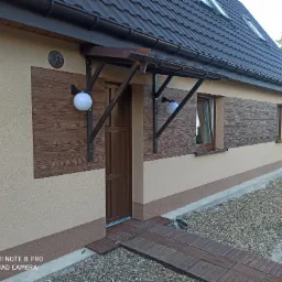 Dom jednorodzinny z elewacją w odcieniach beżu i brązu, z dekoracyjnymi panelami imitującymi drewno, oświetlony kulistymi lampami i zadaszeniem nad wejściem, z drewnianym chodnikiem prowadzącym...