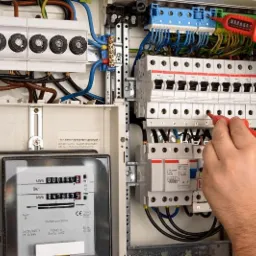 Otwarta skrzynka z bezpiecznikami i licznikiem energii elektrycznej, sprawdzana przez technika za pomocą dwóch mierników napięcia z czerwonymi uchwytami.