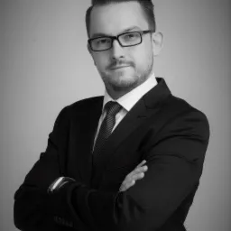 Portret czarno-biały mężczyzny w garniturze, z założonymi rękami, w okularach, z delikatnym uśmiechem, na jasnym tle.