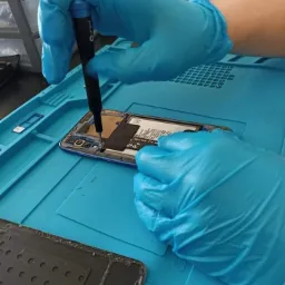 Otwarty smartfon w trakcie naprawy, zabezpieczony niebieską matą, ręce w lateksowych rękawiczkach precyzyjnie operują śrubokrętem, demontując elementy wewnętrzne.