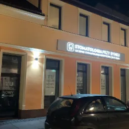 Fasada budynku z szyldem 'Stomatologia Przy Rynku' oświetlona nocą, z zaparkowanymi samochodami na ulicy.