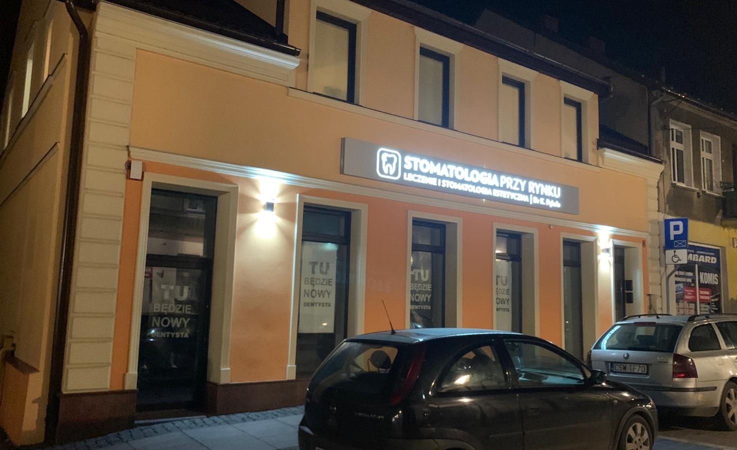 Fasada budynku z szyldem 'Stomatologia Przy Rynku' oświetlona nocą, z zaparkowanymi samochodami na ulicy.