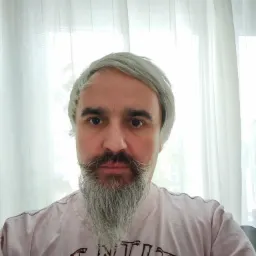 Portret mężczyzny z siwymi włosami i brodą, ubrany w różową koszulkę z nadrukiem, na tle białej, prześwitującej firanki.