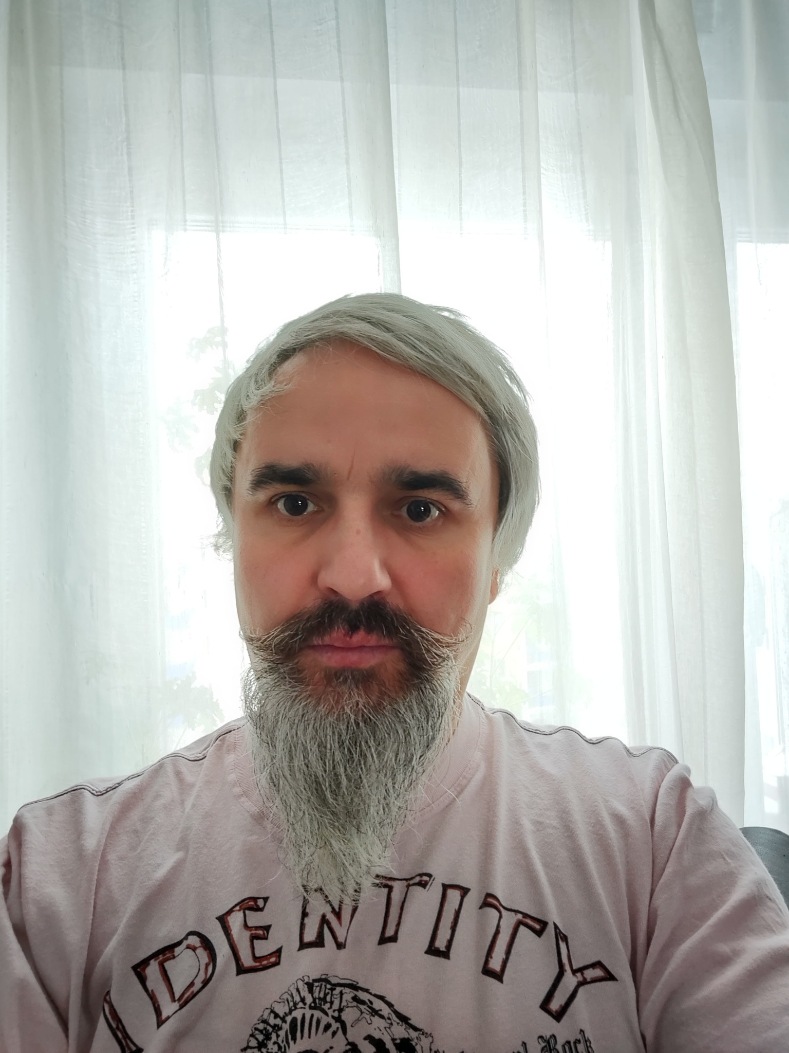 Portret mężczyzny z siwymi włosami i brodą, ubrany w różową koszulkę z nadrukiem, na tle białej, prześwitującej firanki.