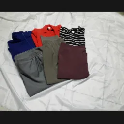 Zestaw ubrań damskich: granatowa bluza z suwakiem, czerwona koszula, szare spodnie, oliwkowe spodnie, czarno-biała bluzka w paski i bordowa spódnica, ułożone na białym, pogniecionym materiale.