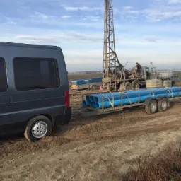 Wiertnica na podwoziu ciężarowym podczas pracy w Łasku, widoczny przyczepiony do samochodu dostawczego transport rur w kolorze niebieskim.