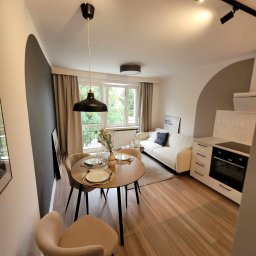 DIM BUD - Jasne mieszkanie po remoncie: drewniana podłoga, okrągły stół z nakryciem, biała sofa z czarną poduszką, aneks kuchenny z piekarnikiem i płytą, czarne lampy sufitowe.