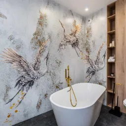 Nowoczesna łazienka z wolnostojącą białą wanną, złotą armaturą, szarą ścianą z motywem żurawi i zabudową meblową z drewnopodobnym wykończeniem