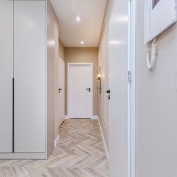 DIM BUD - Nowoczesny korytarz w jasnych barwach z parkietem ułożonym w jodełkę, zabudowaną szafą z długimi, czarnymi uchwytami, białymi drzwiami i minimalistycznym oświetleniem punktowym.