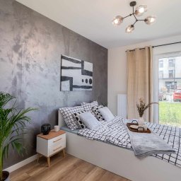 DIM BUD - Sypialnia z łóżkiem w czarno-białą kratę, szarą ścianą z efektem betonu, drewnianą podłogą i rośliną doniczkową w rogu, widok na zewnątrz przez okno balkonowe.