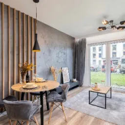 Nowoczesne wnętrze mieszkania z okrągłym stołem jadalnym, szarą ścianą z drewnianymi listwami i dużym oknem balkonowym z widokiem na zewnątrz.