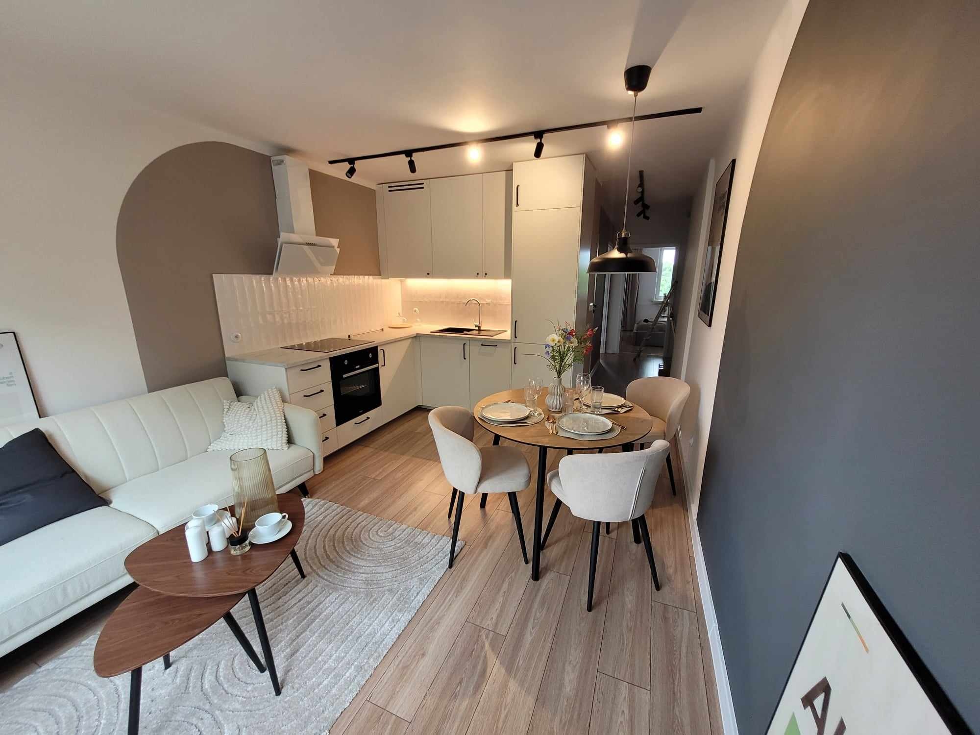 Jasne, nowocześnie urządzone wnętrze mieszkania z aneksem kuchennym, okrągłym stołem jadalnym i sofą w salonie. Widoczne białe szafki kuchenne z oświetleniem punktowym, drewniany stół z nakryciem...