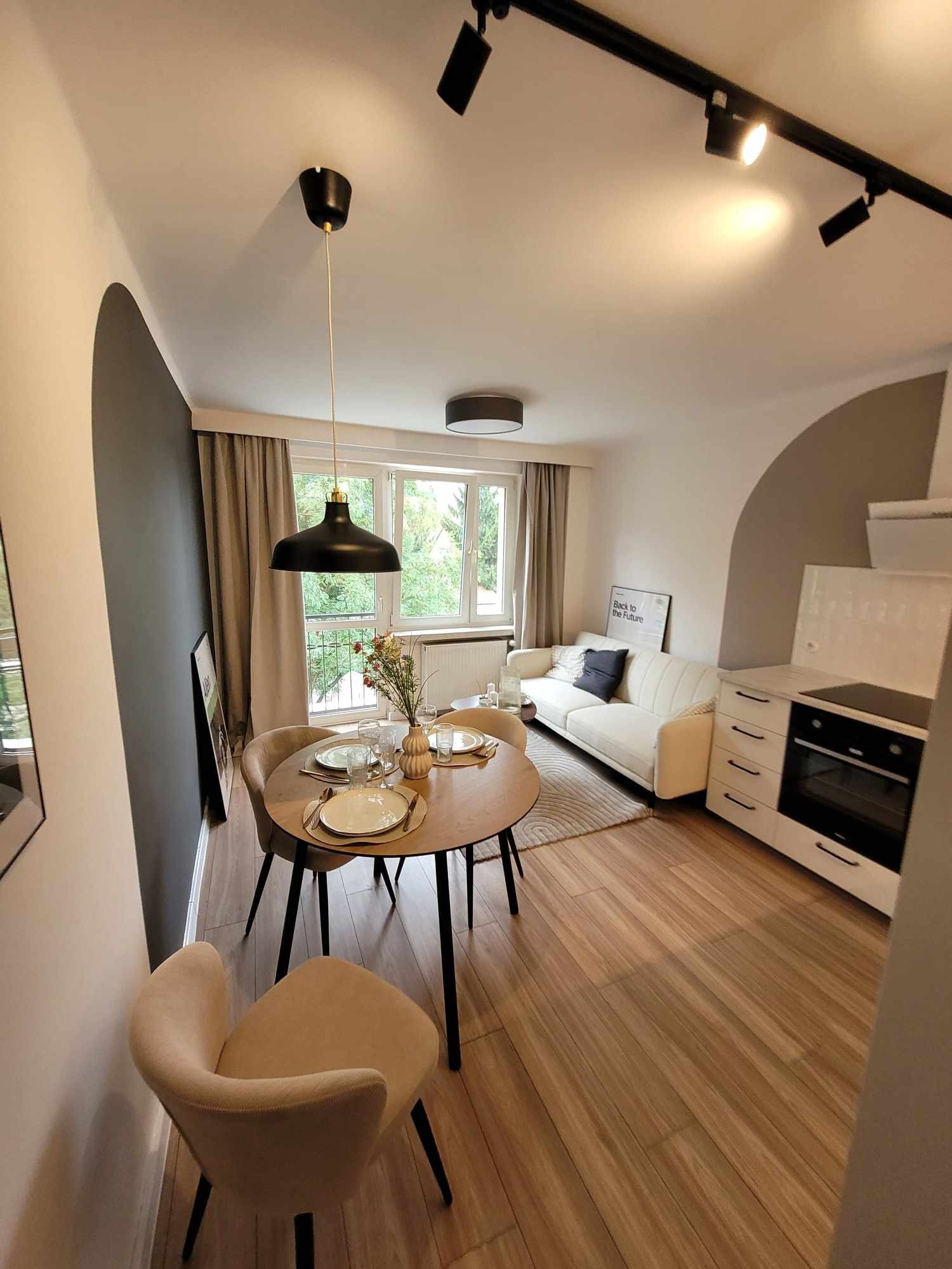Jasne mieszkanie po remoncie: drewniana podłoga, okrągły stół z nakryciem, biała sofa z czarną poduszką, aneks kuchenny z piekarnikiem i płytą, czarne lampy sufitowe.