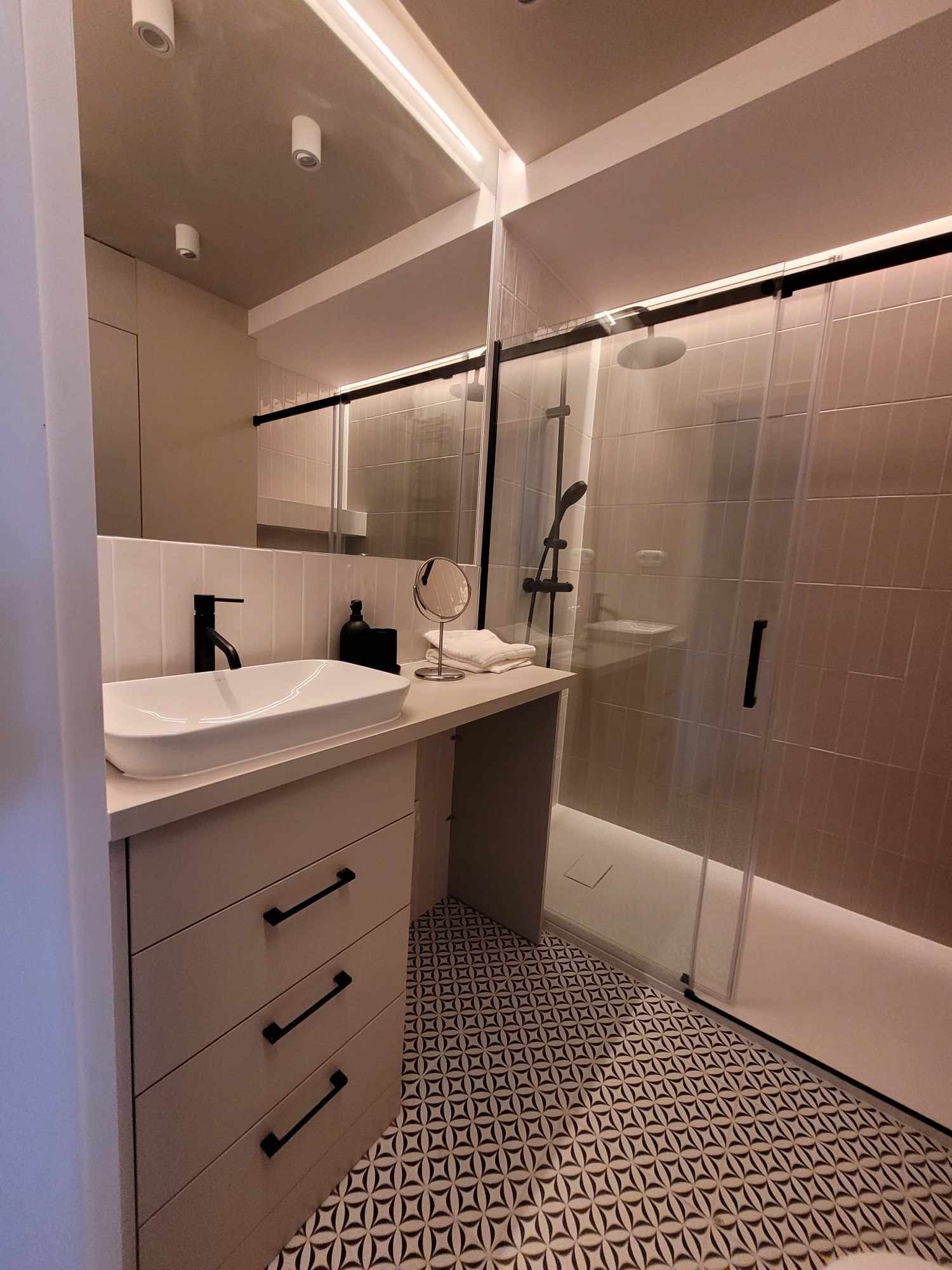 Nowoczesna łazienka z białą umywalką nablatową, czarną armaturą, kabiną prysznicową ze szklanymi drzwiami przesuwnymi i wzorzystą podłogą.