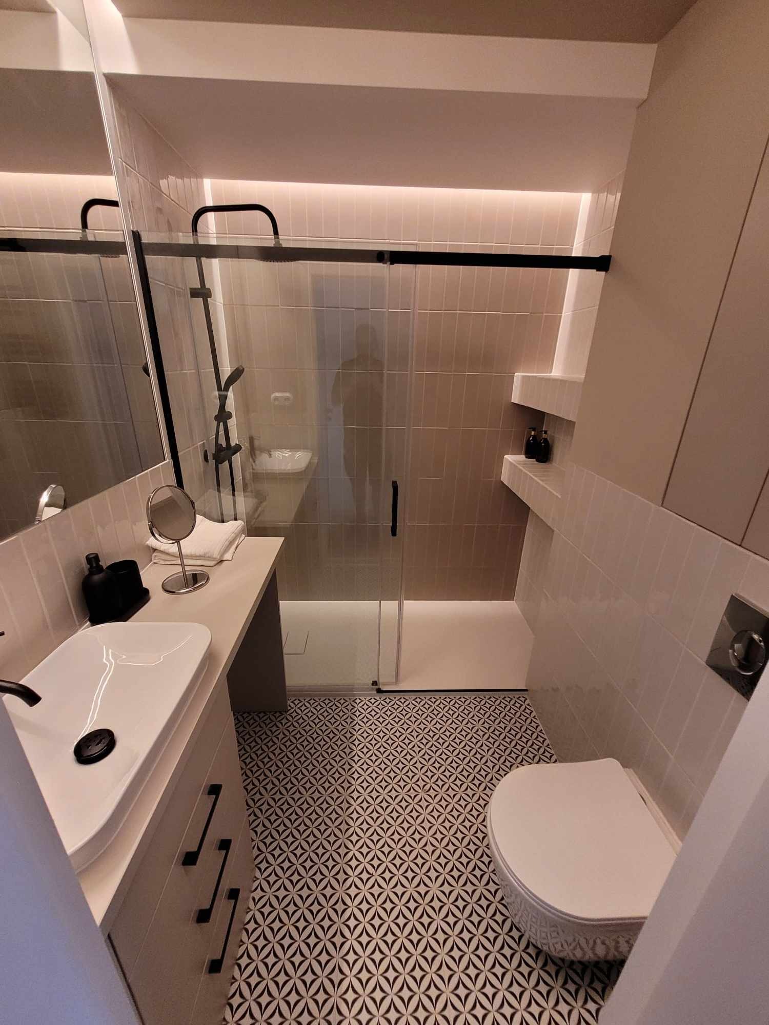 Nowoczesna łazienka z kabiną prysznicową typu walk-in, umywalką nablatową na szafce z czarnymi uchwytami, toaletą i geometryczną posadzką w kolorach czerni i bieli.