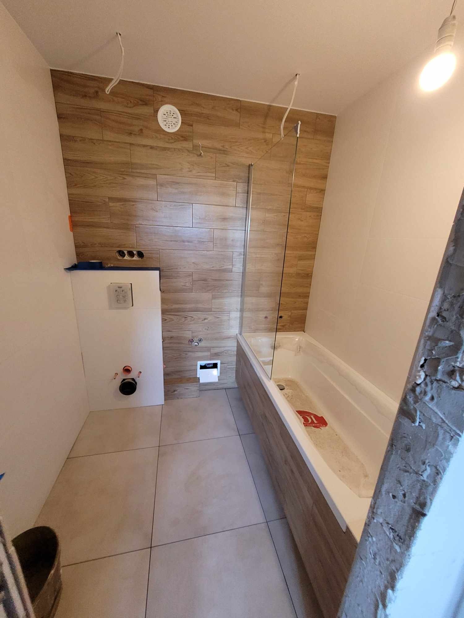 Łazienka w trakcie remontu: drewnopodobne płytki na ścianach, zabudowana wanna z parawanem, podwieszany sedes, widoczne kable oświetleniowe.