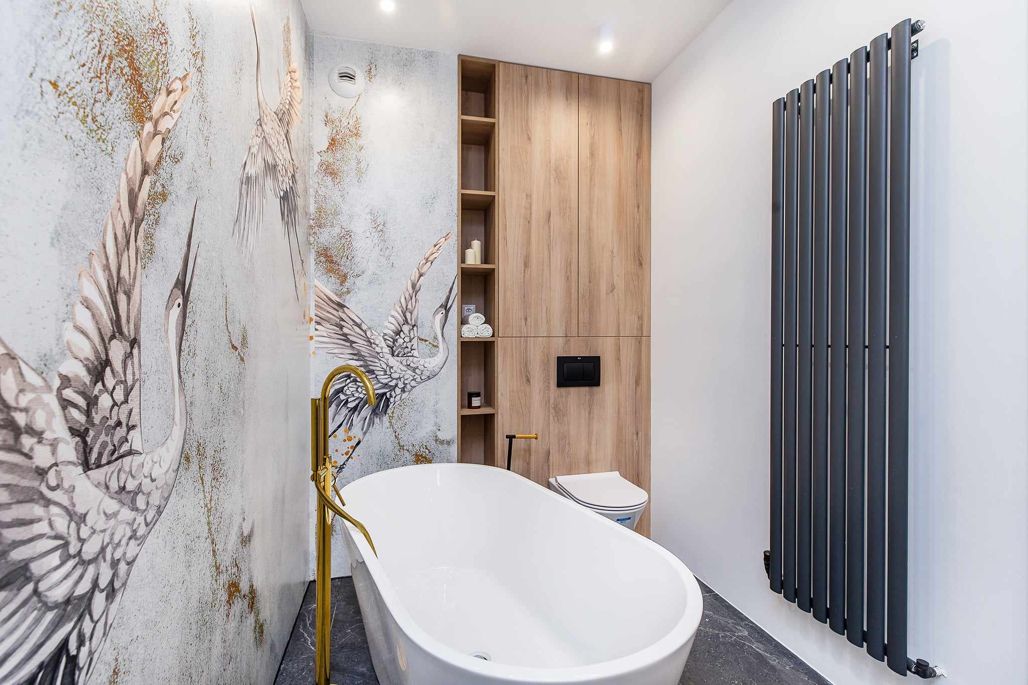 Nowoczesna łazienka w Tłuczani z wolnostojącą białą wanną, złotą armaturą, tapetą z motywem żurawi, drewnianą zabudową i grafitowym grzejnikiem.
