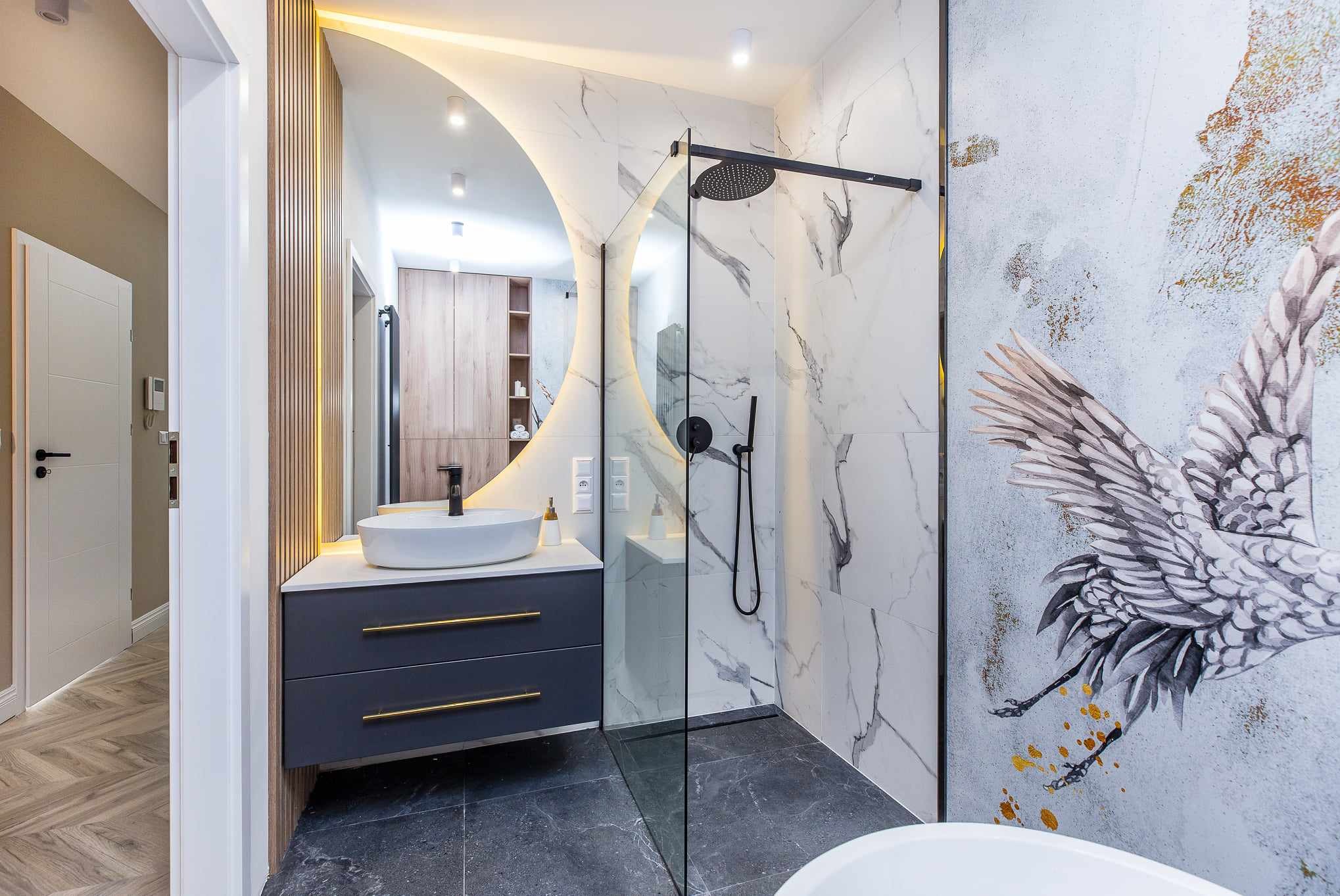 Nowoczesna łazienka z kabiną prysznicową typu walk-in, umywalką nablatową na szafce z szufladami z złotymi uchwytami, okrągłym lustrem z podświetleniem LED i dekoracyjną tapetą z motywem żurawia.