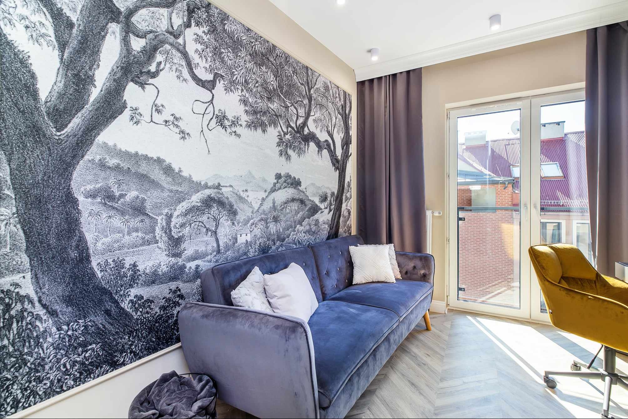 Salon z fototapetą przedstawiającą krajobraz, niebieska sofa z poduszkami i widok na zewnątrz przez okno balkonowe.