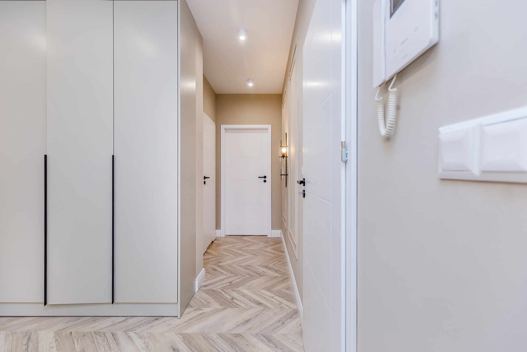 Nowoczesny korytarz w jasnych barwach z parkietem ułożonym w jodełkę, zabudowaną szafą z długimi, czarnymi uchwytami, białymi drzwiami i minimalistycznym oświetleniem punktowym.