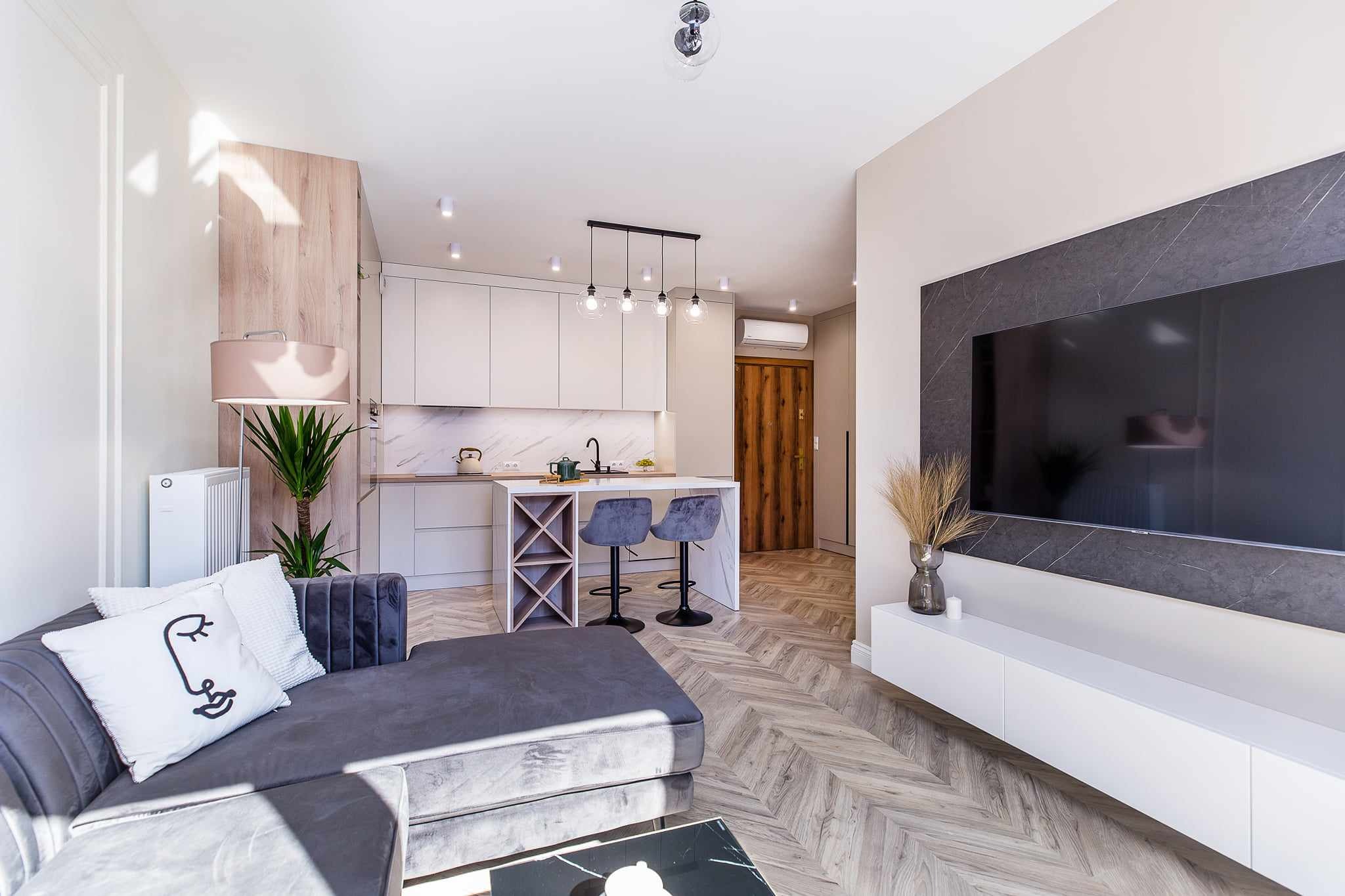 Nowoczesne wnętrze mieszkania z otwartą kuchnią, szarą sofą z poduszką z motywem twarzy, parkietem w jodełkę i telewizorem z marmurową ramą.