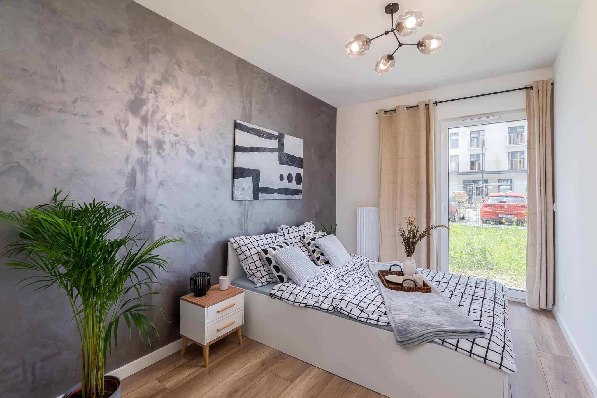 Sypialnia z łóżkiem w czarno-białą kratę, szarą ścianą z efektem betonu, drewnianą podłogą i rośliną doniczkową w rogu, widok na zewnątrz przez okno balkonowe.
