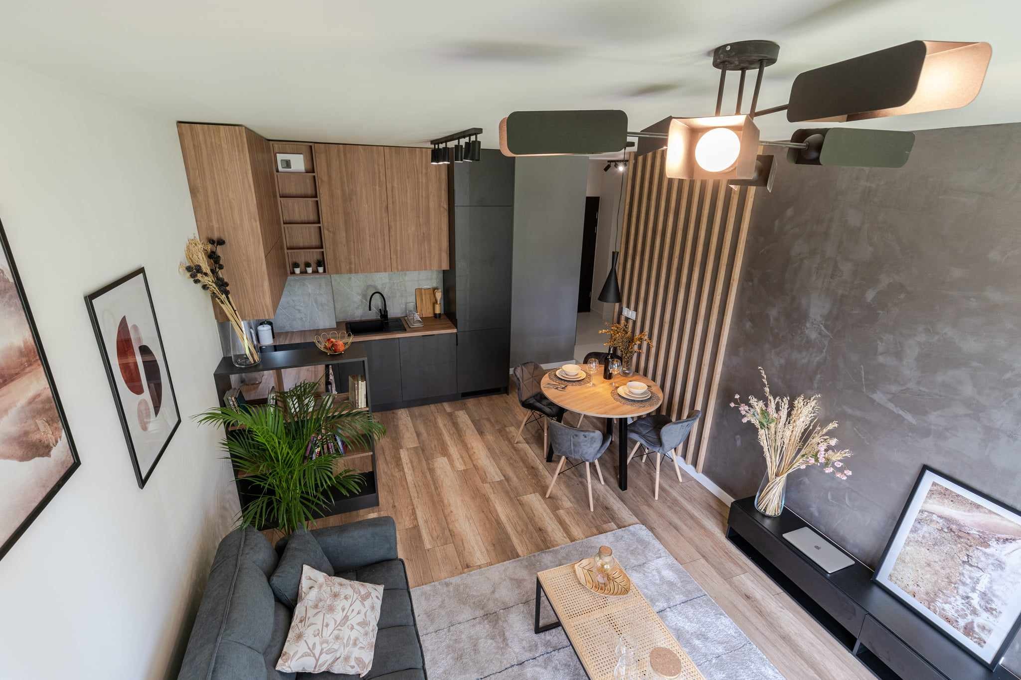 Widok z góry na nowocześnie urządzone wnętrze mieszkania z drewnianą podłogą, szarą sofą z wzorzystą poduszką, okrągłym stołem jadalnym i aneksem kuchennym z drewnianymi szafkami.