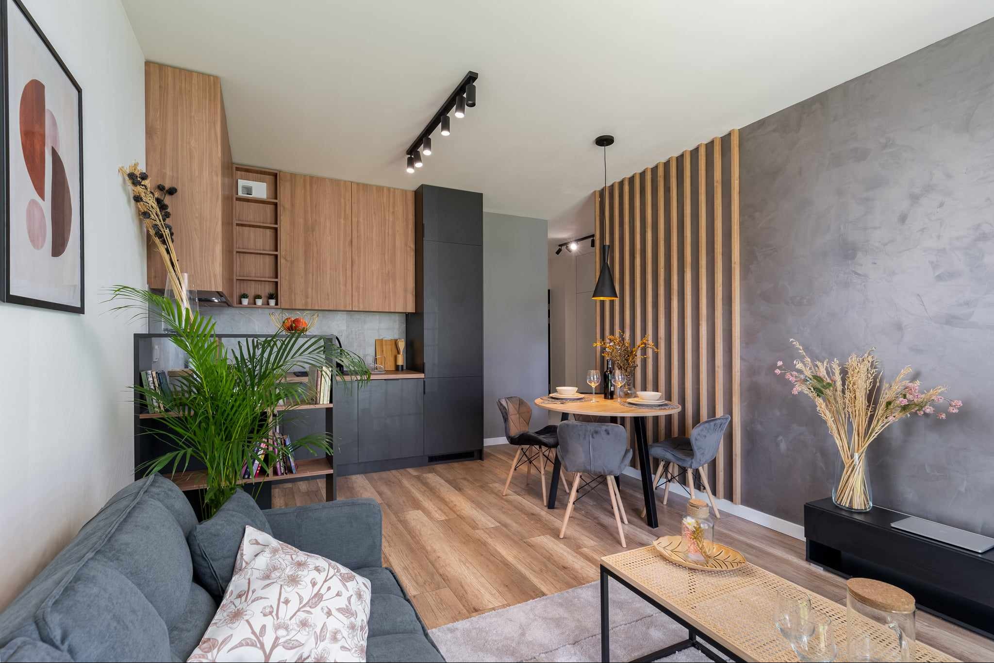 Nowoczesne wnętrze salonu z aneksem kuchennym: szara sofa z poduszką w kwiaty, drewniana podłoga, stół z krzesłami, kuchnia z drewnianymi i grafitowymi szafkami, ściana z dekoracyjnymi pionowymi...