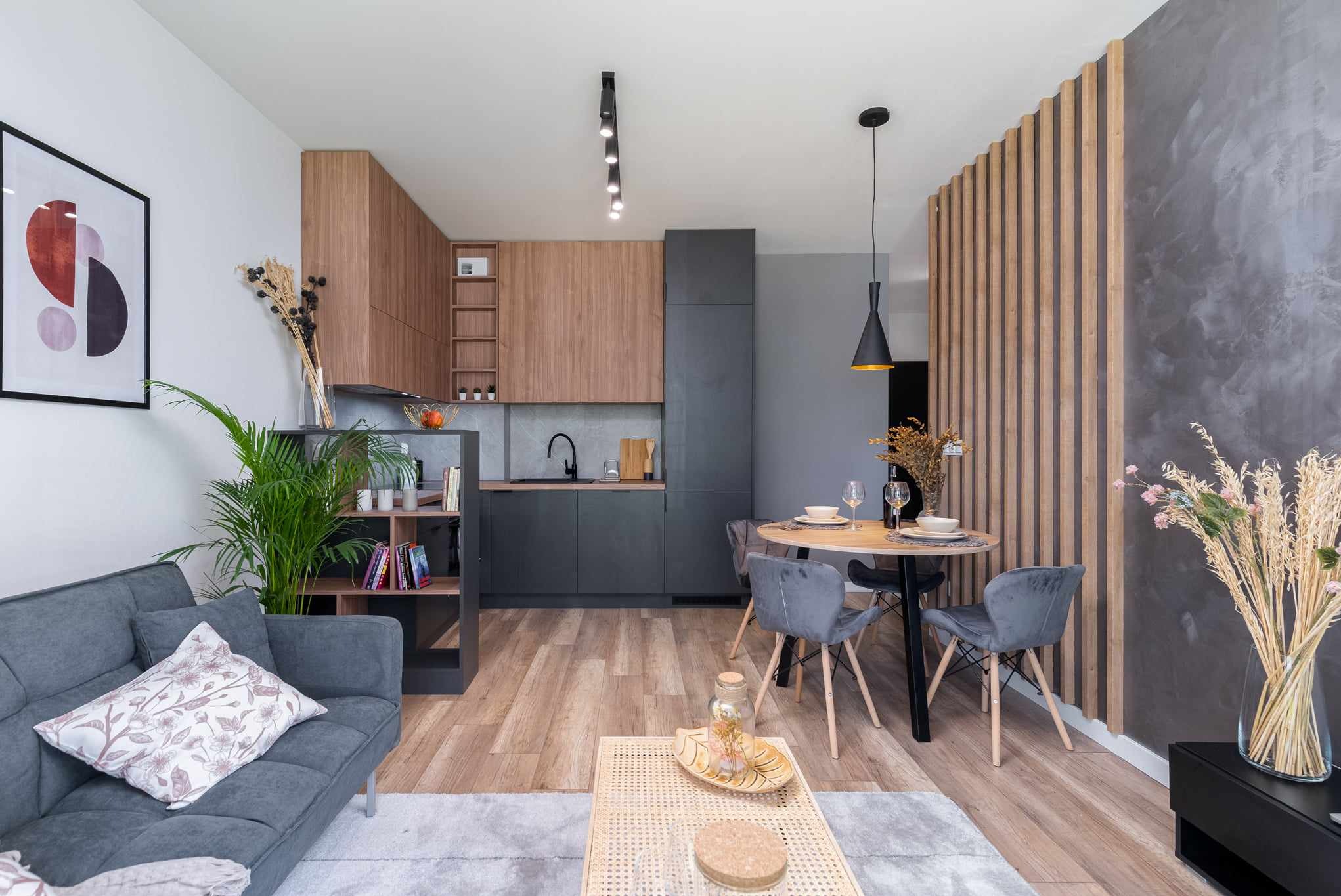 Nowoczesne wnętrze salonu z aneksem kuchennym, szara sofa z wzorzystą poduszką, drewniane panele dekoracyjne na ścianie, okrągły stół z szarymi krzesłami, drewniane i grafitowe szafki kuchenne...