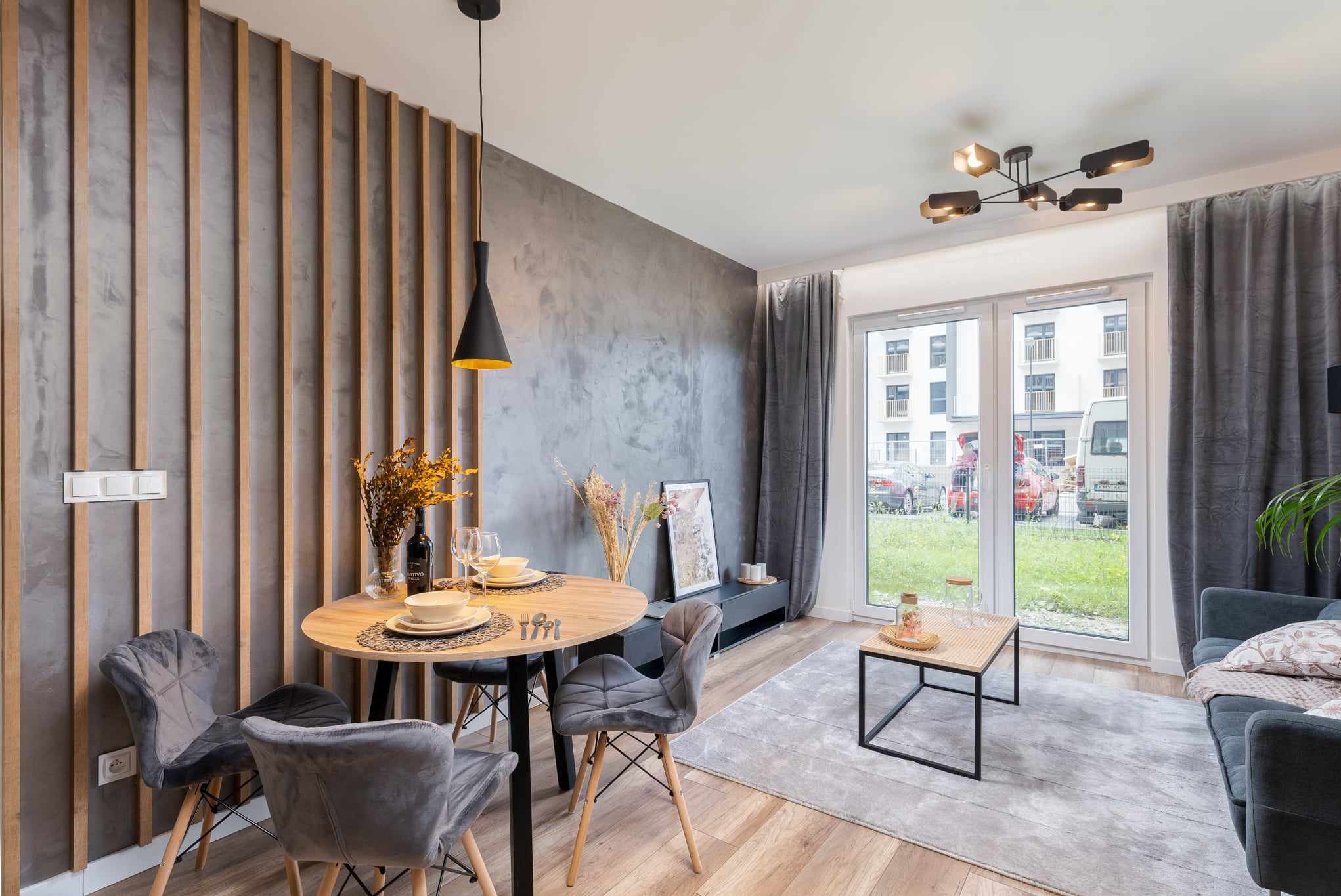 Nowoczesne wnętrze mieszkania z okrągłym stołem jadalnym, szarą ścianą z drewnianymi listwami i dużym oknem balkonowym z widokiem na zewnątrz.