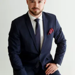 Mężczyzna z brodą w granatowym garniturze, białej koszuli i ciemnym krawacie, siedzący na stołku, z ozdobną poszetką w kieszonce marynarki i zegarkiem na nadgarstku.