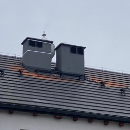 INTERBUD-MD Sp.z.o.o. - Dwa szare kominy z metalowymi daszkami na ciemnym, spadzistym dachu z dachówki ceramicznej, widoczne elementy systemu przeciwśniegowego, fragment ściany budynku z oknem w tle.