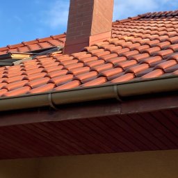INTERBUD-MD Sp.z.o.o. - Czerwona dachówka ceramiczna na dachu domu z ceglanym kominem, widoczne brązowe rynny i fragment okna dachowego.
