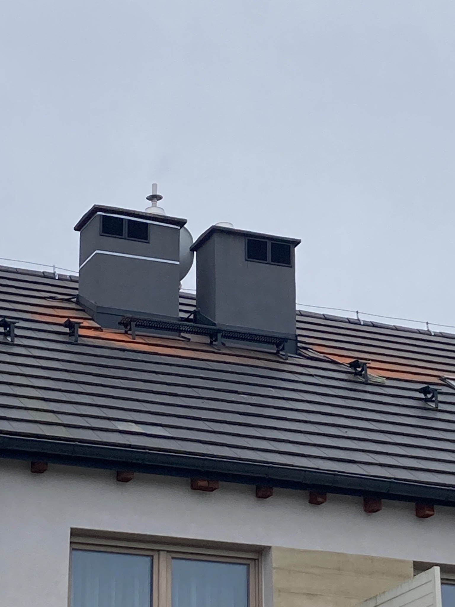 Dwa szare kominy z metalowymi daszkami na ciemnym, spadzistym dachu z dachówki ceramicznej, widoczne elementy systemu przeciwśniegowego, fragment ściany budynku z oknem w tle.