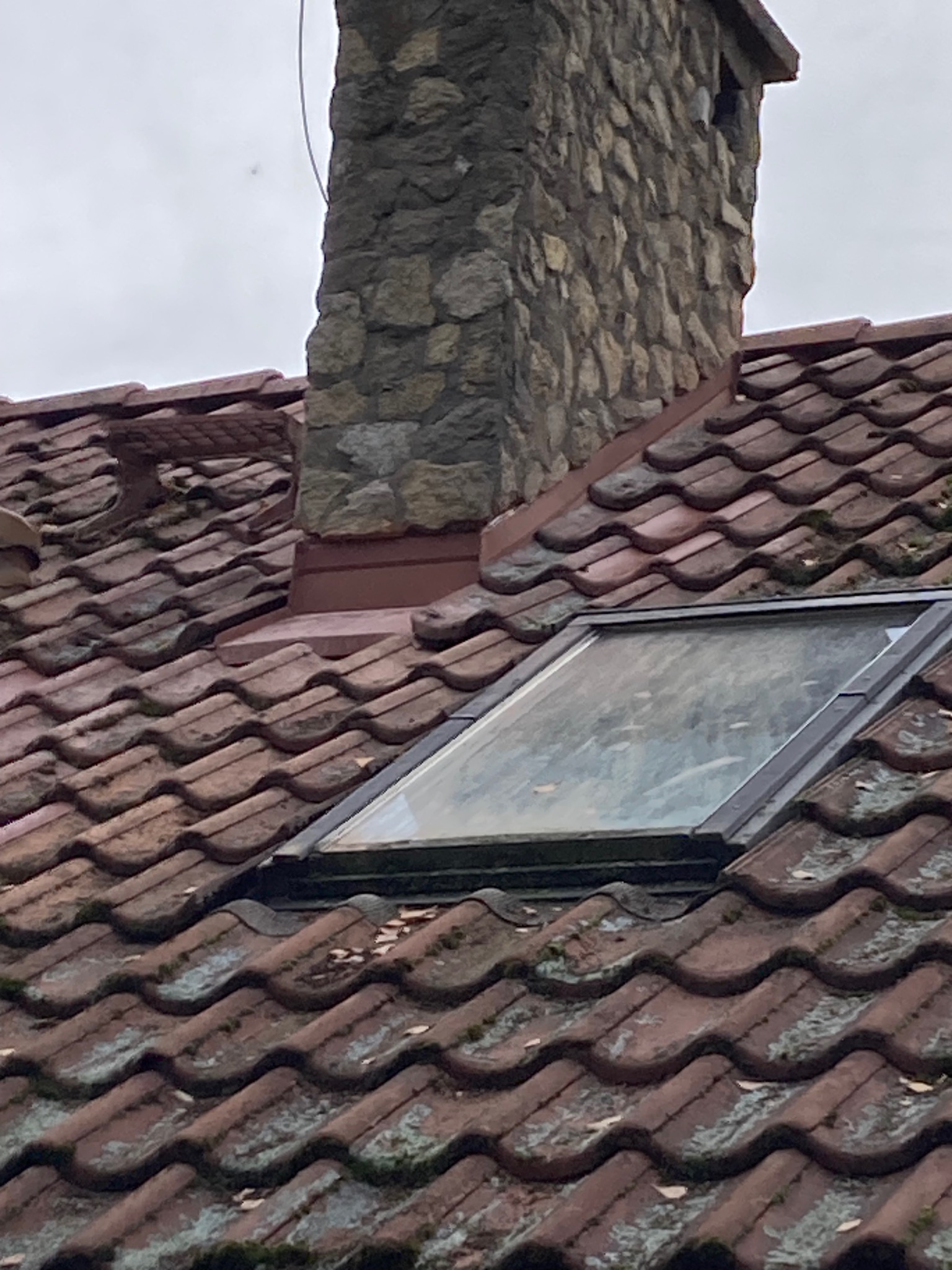 Czerwona dachówka ceramiczna z oknem dachowym i kamiennym kominem, widoczne ślady mchu i zabrudzeń.