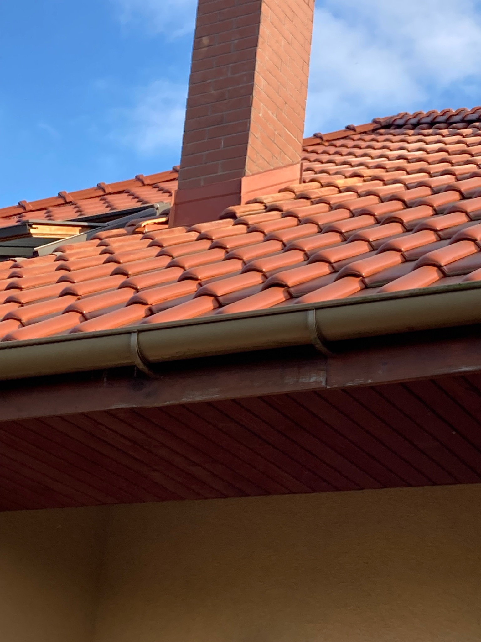 Czerwona dachówka ceramiczna na dachu domu z ceglanym kominem, widoczne brązowe rynny i fragment okna dachowego.
