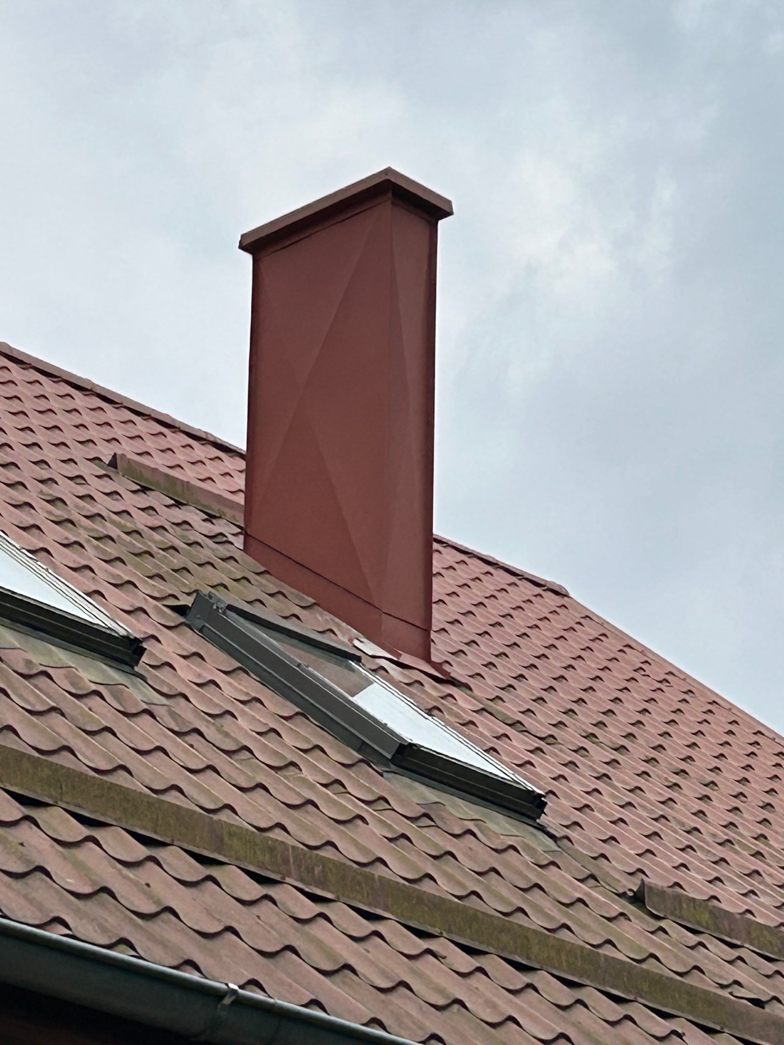 Czerwony dach z dachówką ceramiczną, dwa okna dachowe i wysoki, kwadratowy komin w kolorze dachu na tle pochmurnego nieba.