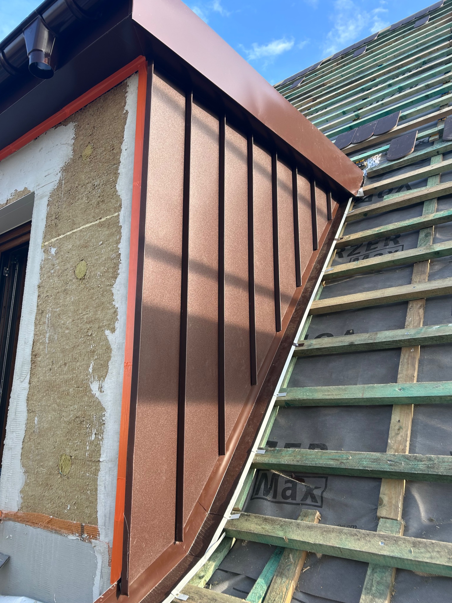 Montaż brązowej blachy na lukarnie, widoczne ocieplenie ściany szczytowej i fragment konstrukcji dachu z membraną, w tle błękitne niebo.