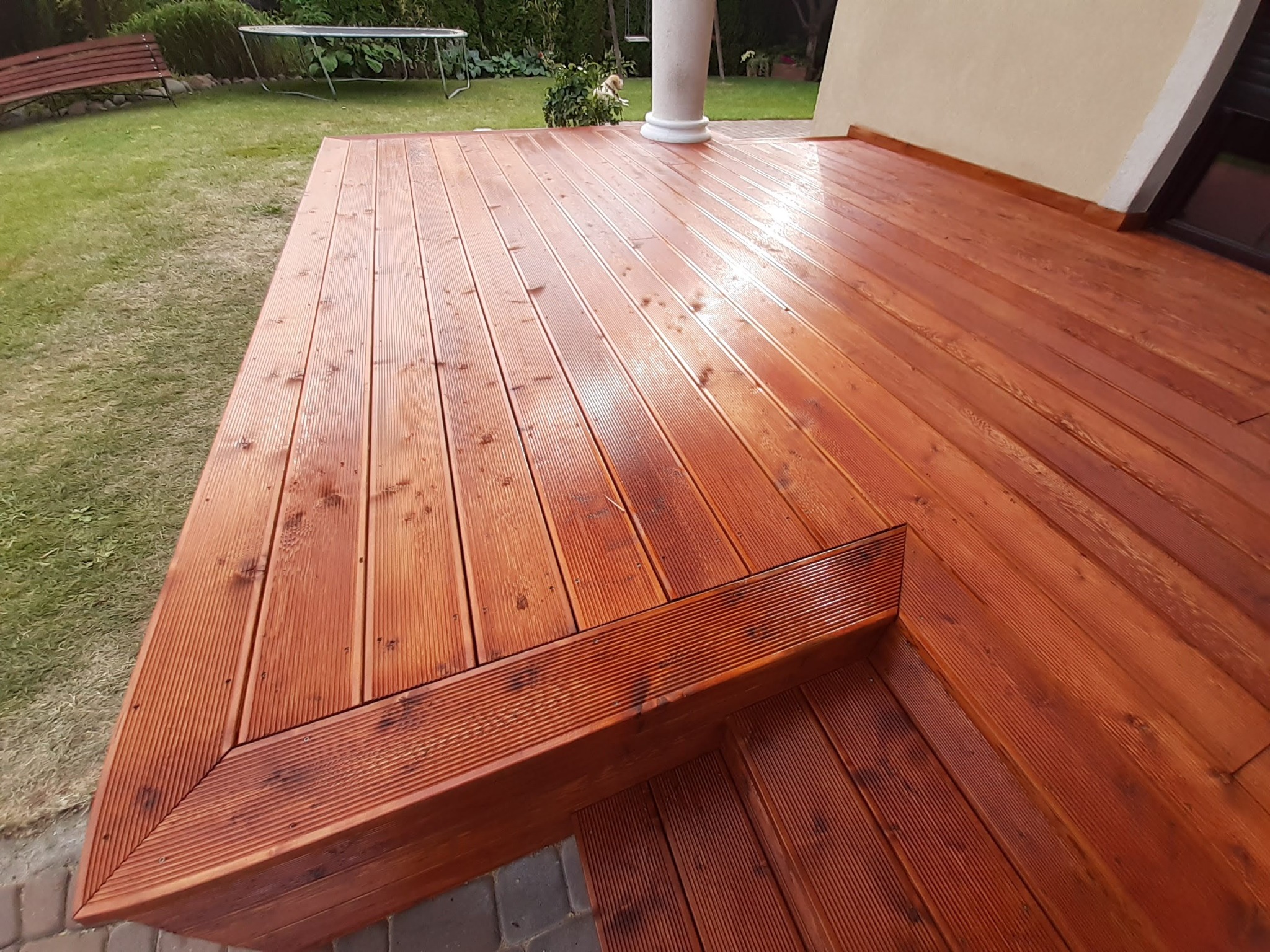 Świeżo polakierowany taras z drewna modrzewiowego w kolorze teak, z niskimi schodkami, otoczony zielonym trawnikiem i elementami architektury ogrodowej.