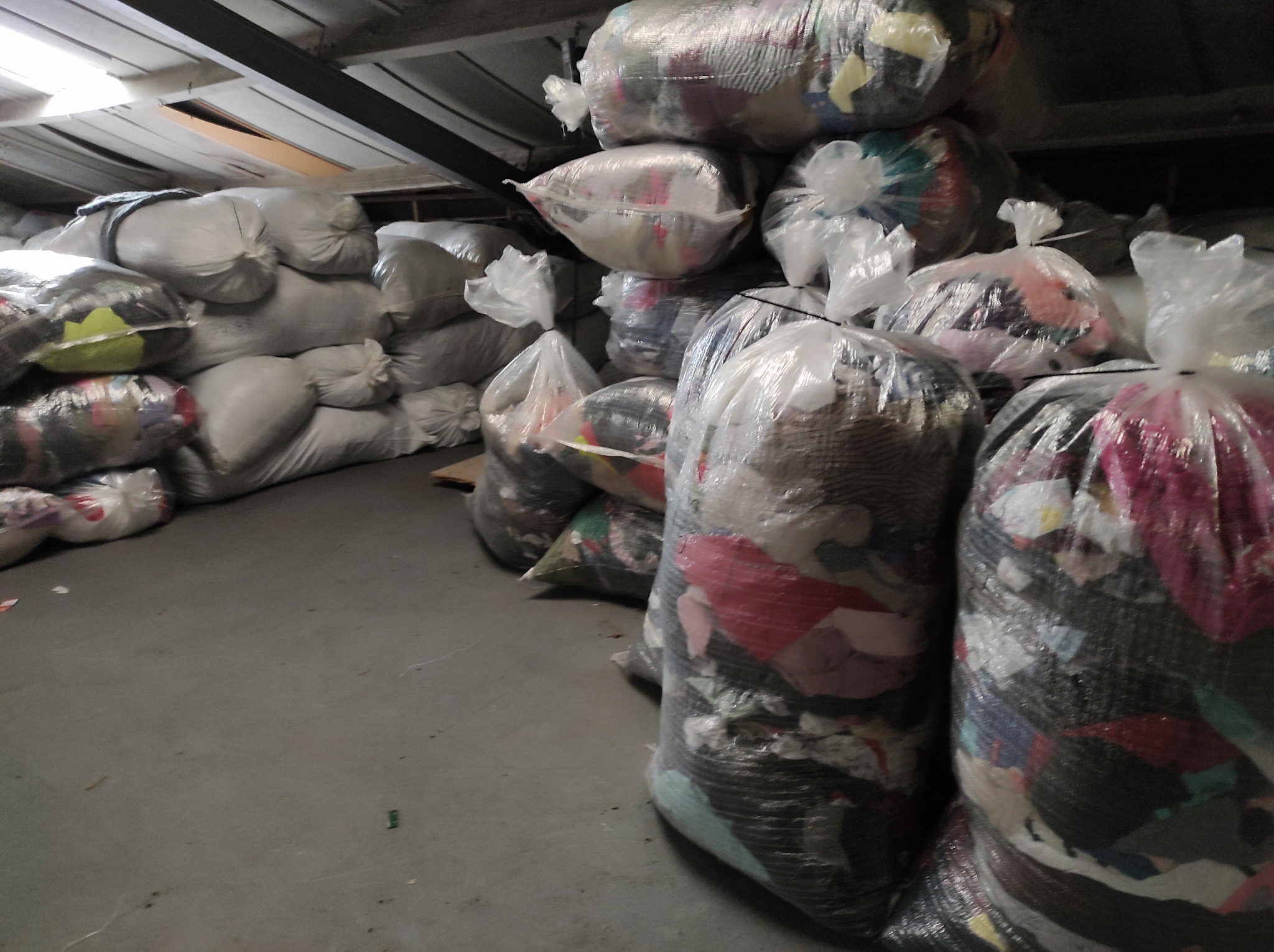 Stos worków z odzieżą, niektóre przezroczyste, ukazujące zawartość, ustawione w magazynie pod skośnym dachem.
