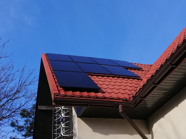 Panele słoneczne na dachu domu z czerwoną dachówką, widoczne rynny i antena telewizyjna w tle na tle błękitnego nieba.